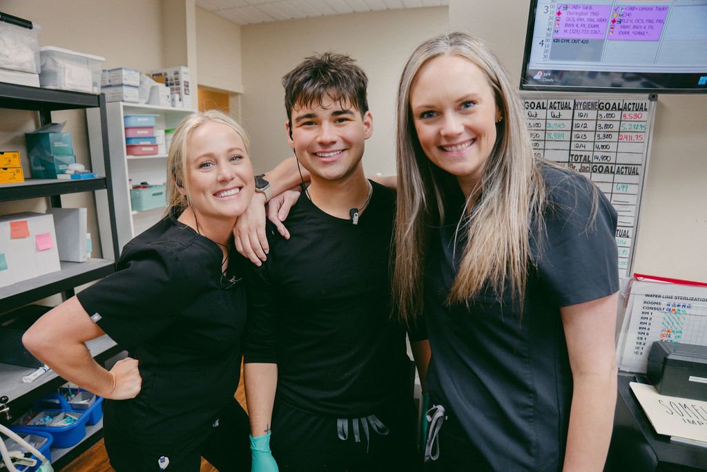 Three smiling healthcare workers in scrubs standing together in a medical office, with shelves and a monitor displaying schedules and goals in the background.