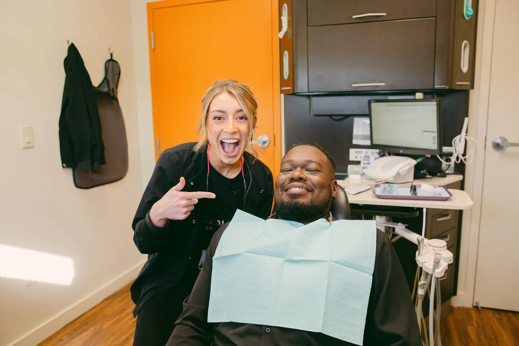 A woman in medical scrubs pointing and smiling next to a man in a dental chair with a bib, inside a dental office.