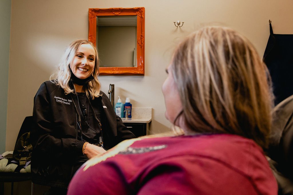 A smiling female dental professional in a black jacket sitting across from a patient in a dental office.