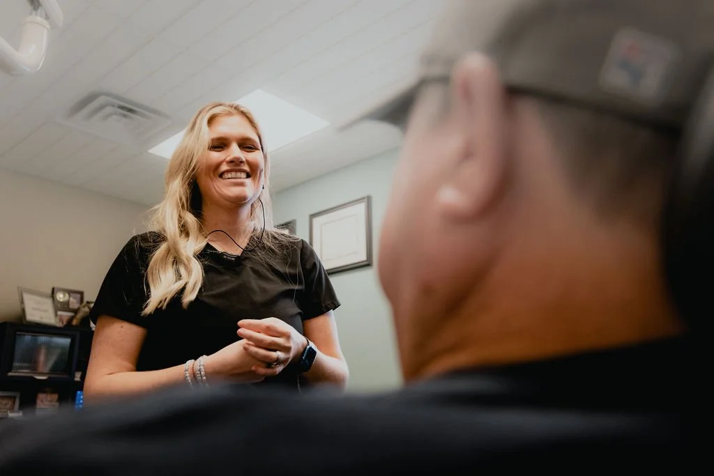 A woman smiling and laughing, wearing a black shirt, standing indoors with a ceiling light, framed certificates on the wall, and a TV or computer screen in the background, viewed from behind a person with short hair and glasses.