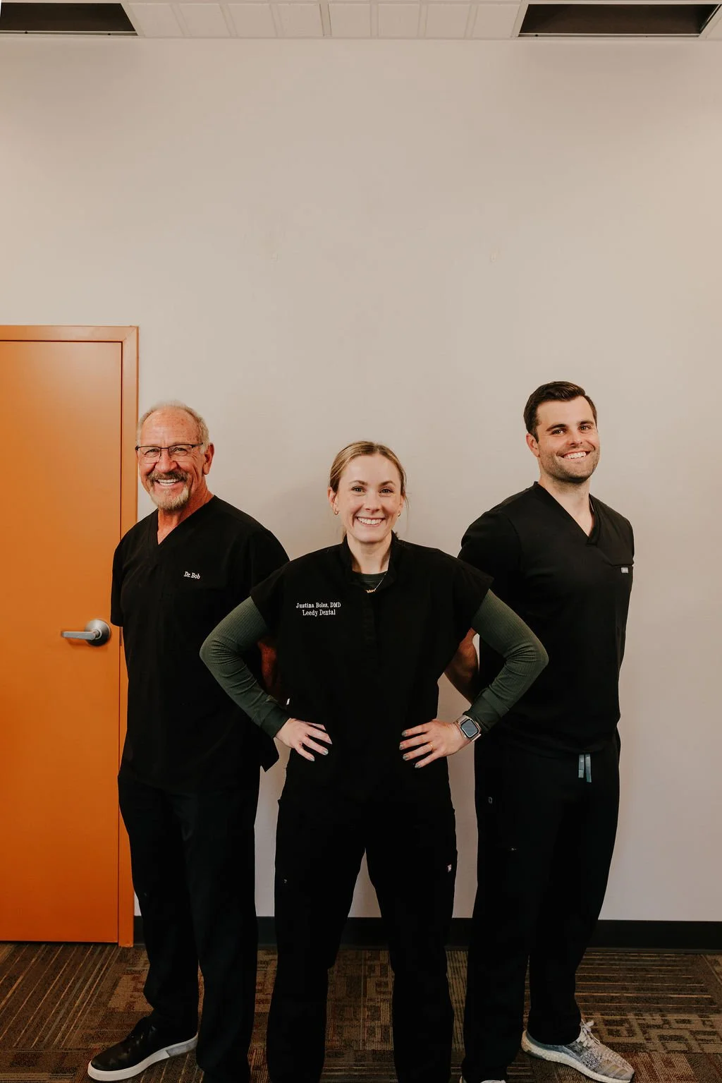 Three dental professionals standing together in an indoor setting, smiling, with a white wall and orange door in the background.