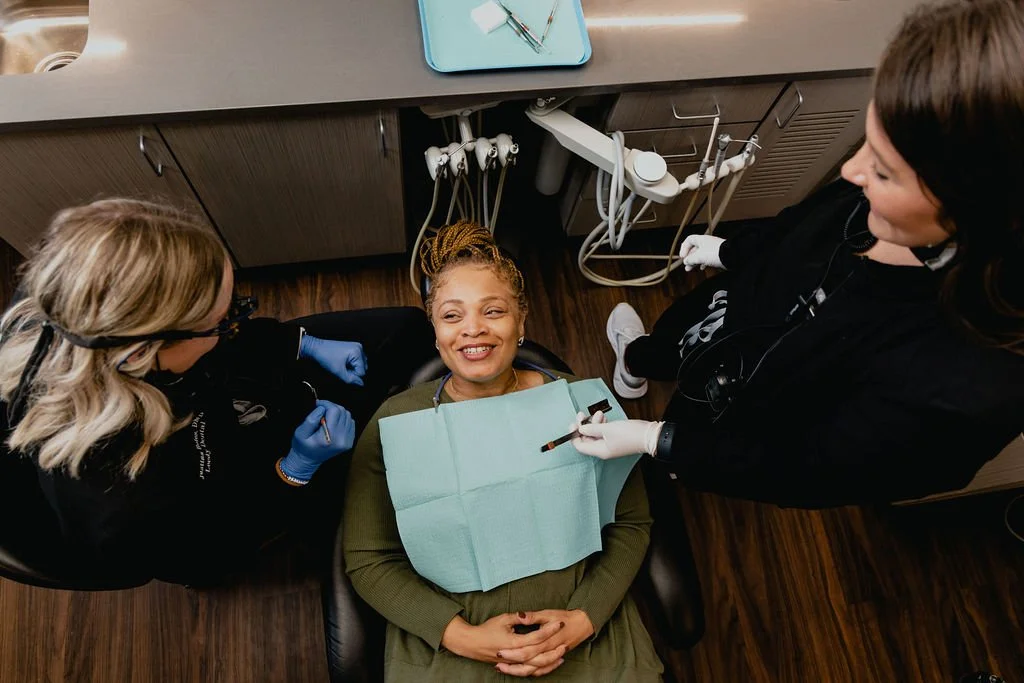 A woman receives dental treatment at a dental clinic, lying back in the dental chair wearing a bib, while two dental hygienists attend to her, one holding dental tools and the other preparing equipment.