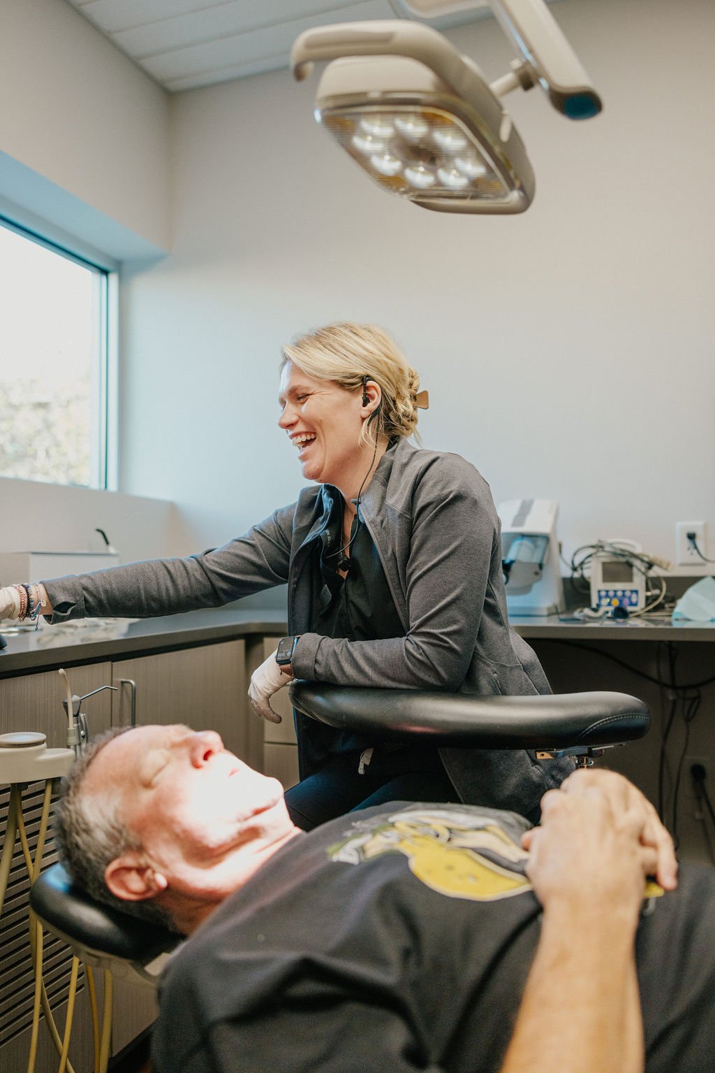 A woman and a man in a medical setting, with the woman smiling and interacting with the man who is lying down, possibly during a dental or medical procedure, in a clinical room with equipment and a window.