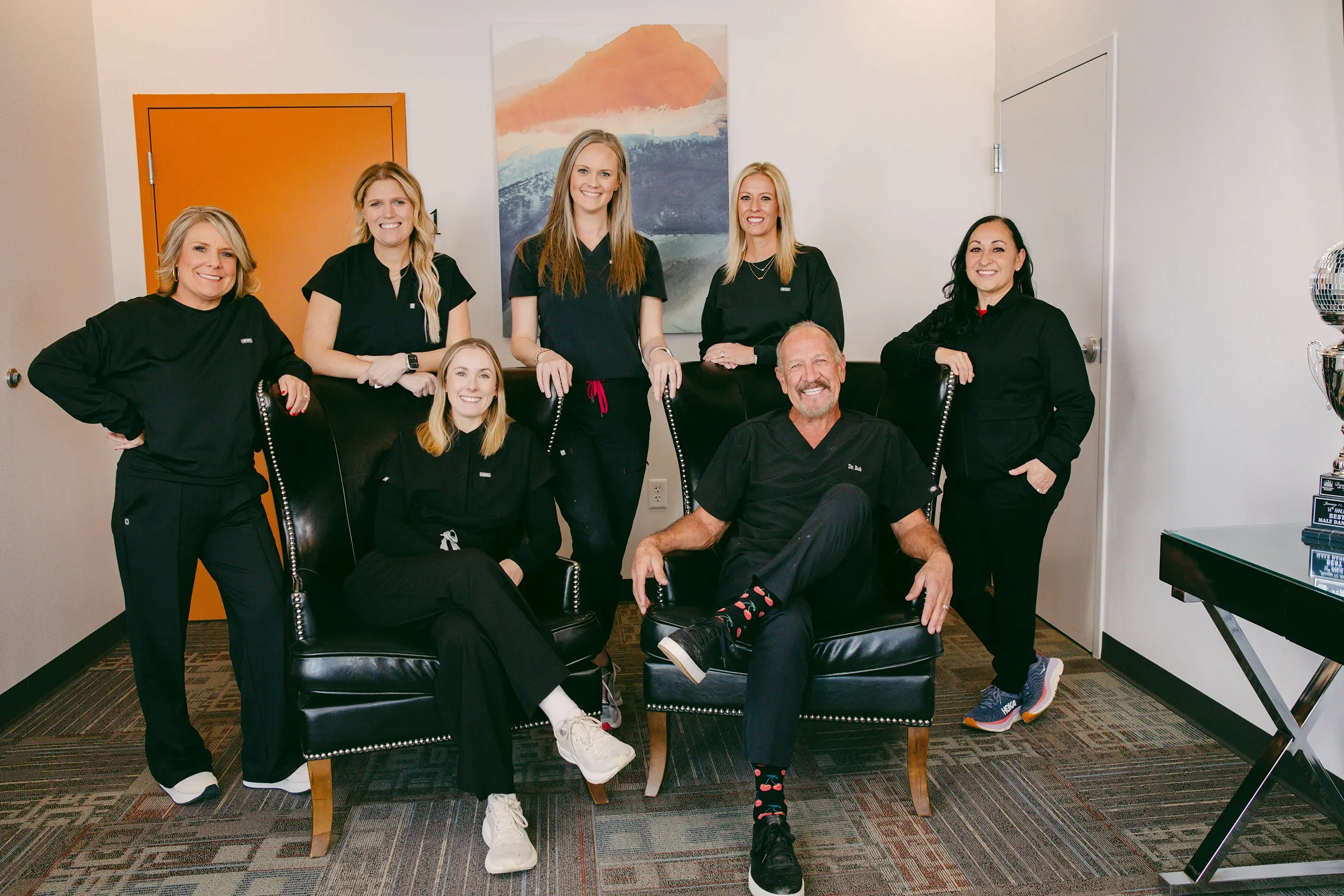 A group of eight people, including one man sitting in a black armchair and seven women standing or sitting around him, posing for a photo inside a room with white walls and modern decor.