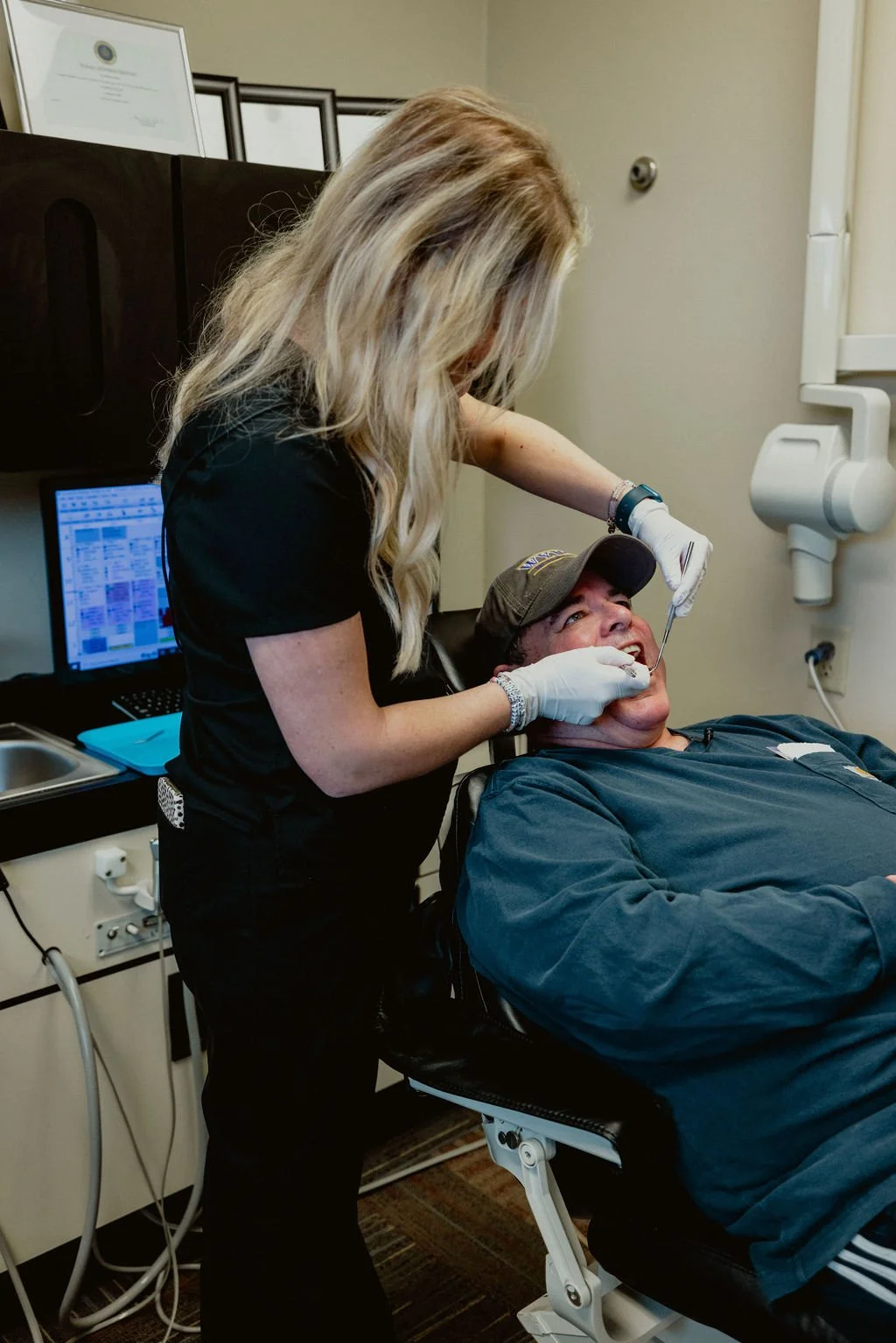 A dentist or dental hygienist performing a dental procedure on a male patient lying in a dental chair, with dental tools and equipment visible in the room.