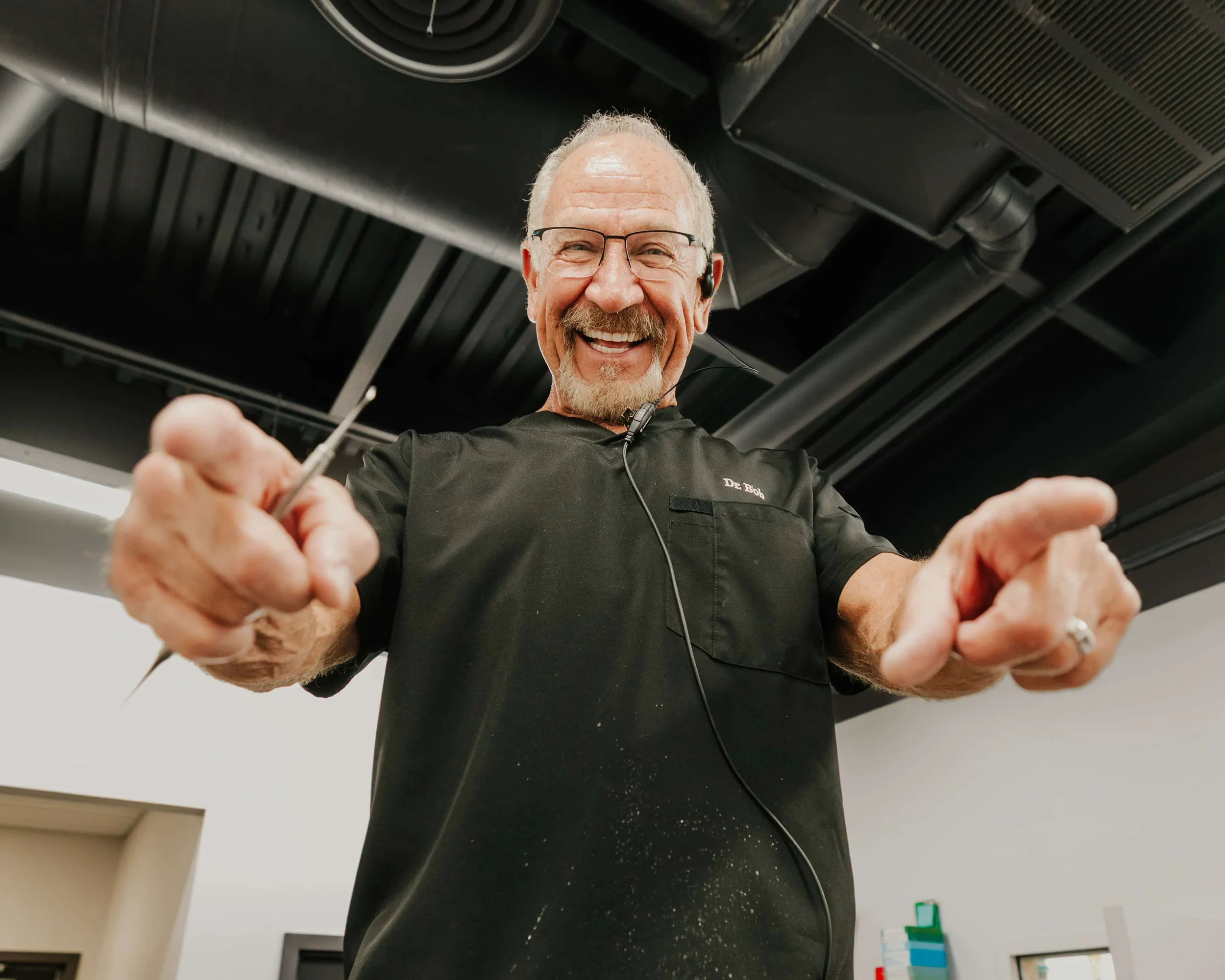Smiling older man with glasses and gray hair, wearing a black shirt with a name tag that says 'Dr. Bob', holding a pair of tweezers, in a room with a black ceiling and white walls.