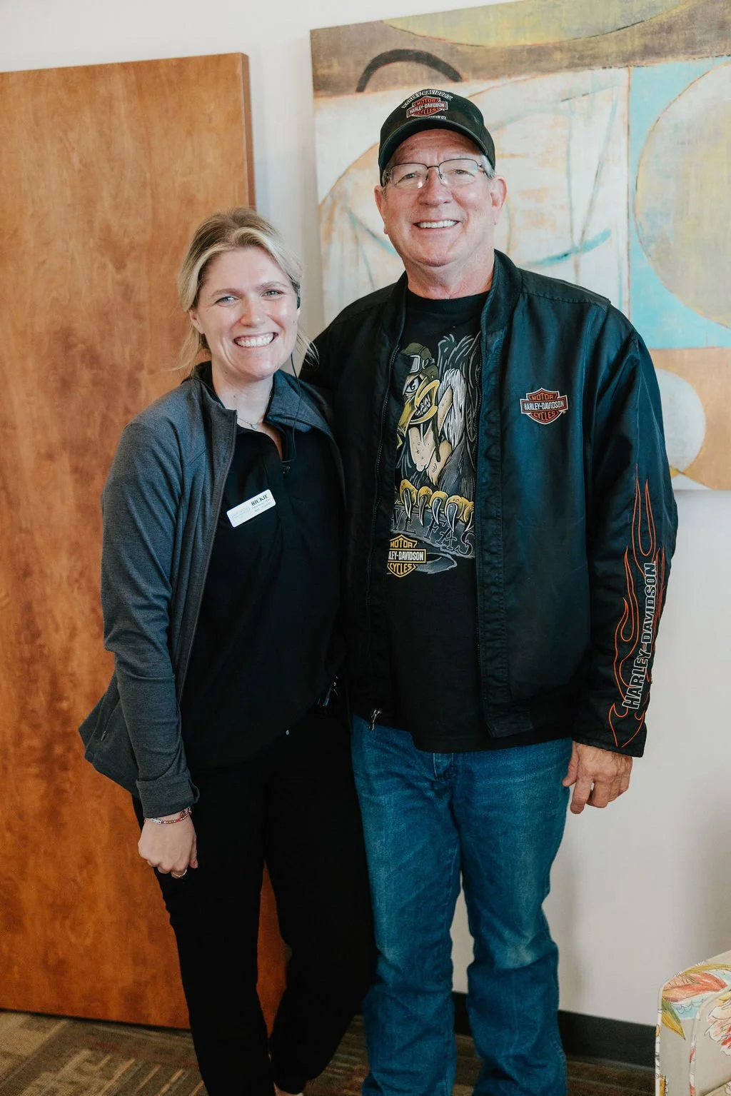 A woman and a man standing side by side indoors, smiling at the camera. The woman is wearing a black shirt with a gray jacket and has a name tag that says 'RACHEL.' The man is wearing a Harley-Davidson jacket, a graphic t-shirt, a black cap, and glasses. They are in front of a wooden door and a colorful abstract painting.