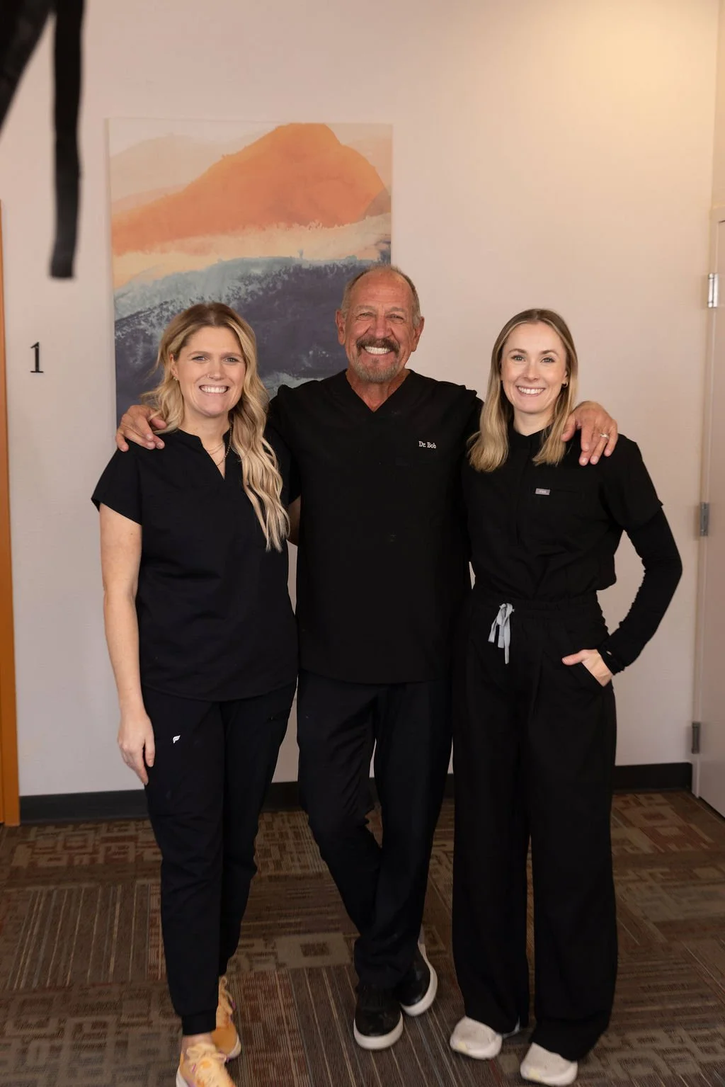 Three smiling healthcare professionals in black scrubs standing arm-in-arm indoors with a colorful abstract wall art in the background.