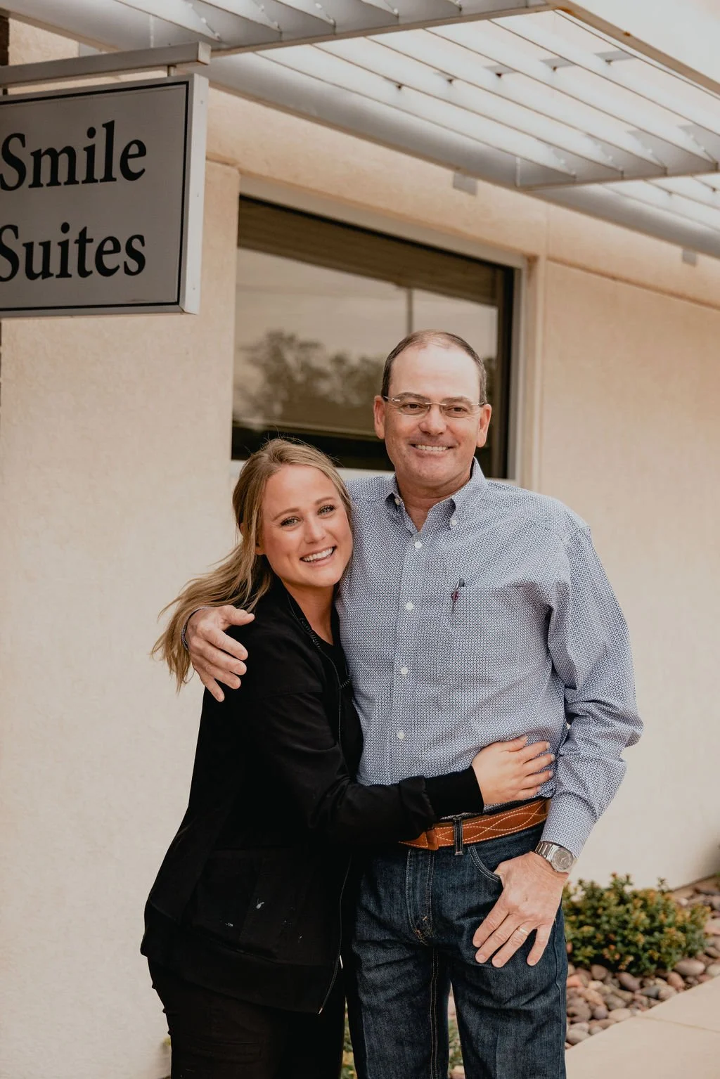 A smiling woman and man standing outside near a sign that reads 'Smile Suites'. The woman has blond hair and is wearing a black jacket, and the man has glasses and is wearing a light blue button-up shirt and jeans.