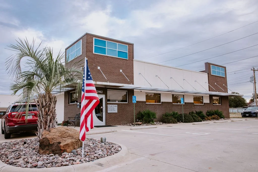 A two-story brick and white building with large windows, a parking lot with wheelchair accessible spots, and an American flag in front. There is a palm tree and some rocks in the landscaped area.