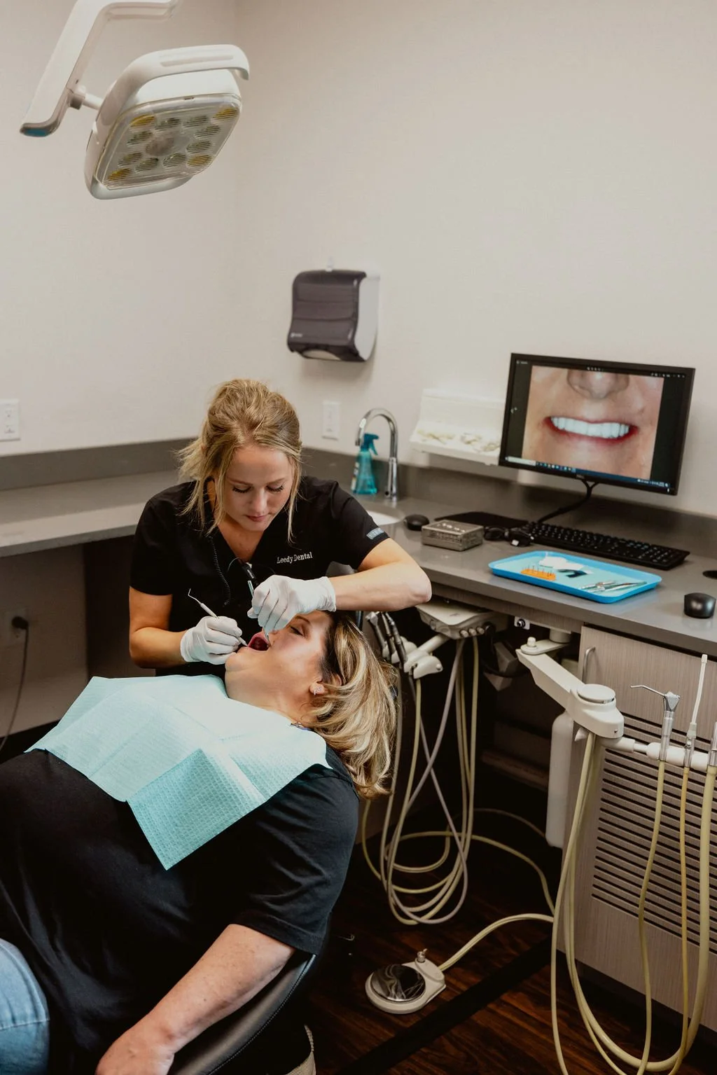 A woman lying in a dental chair receiving dental care from a dentist in a dental office. There is dental equipment, a computer monitor showing a smiling mouth, and dental tools present.