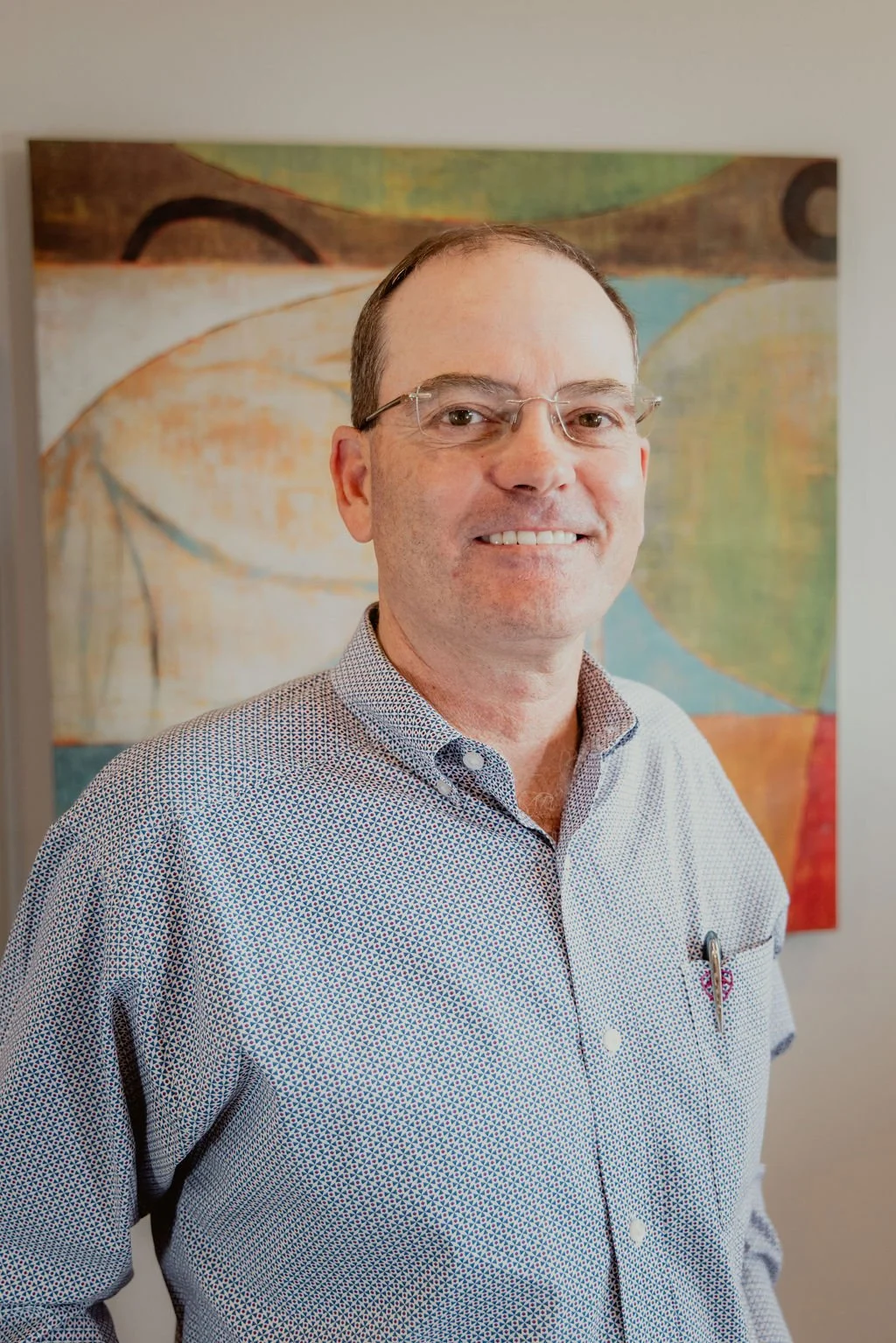 A man wearing glasses and a patterned button-up shirt standing in front of an abstract colorful painting.