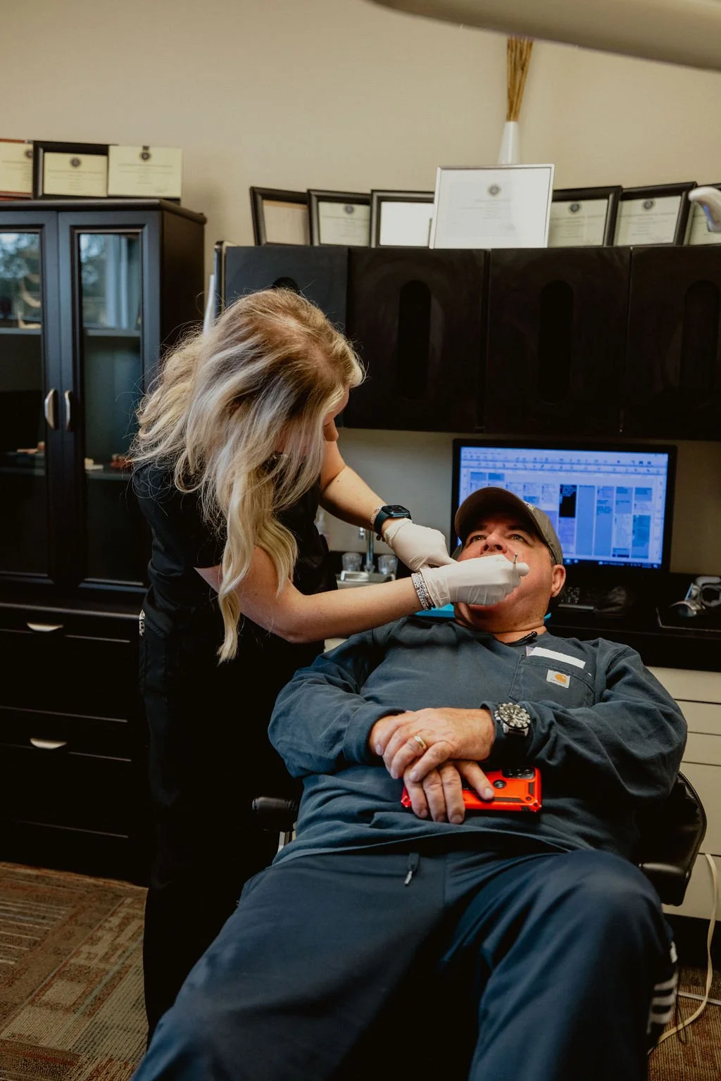 A woman is administering a nasal swab test to a man sitting in a chair. The man is looking up and has his arms crossed, holding a small orange device. The room has a computer with a screen displaying charts and multiple framed certificates or documen