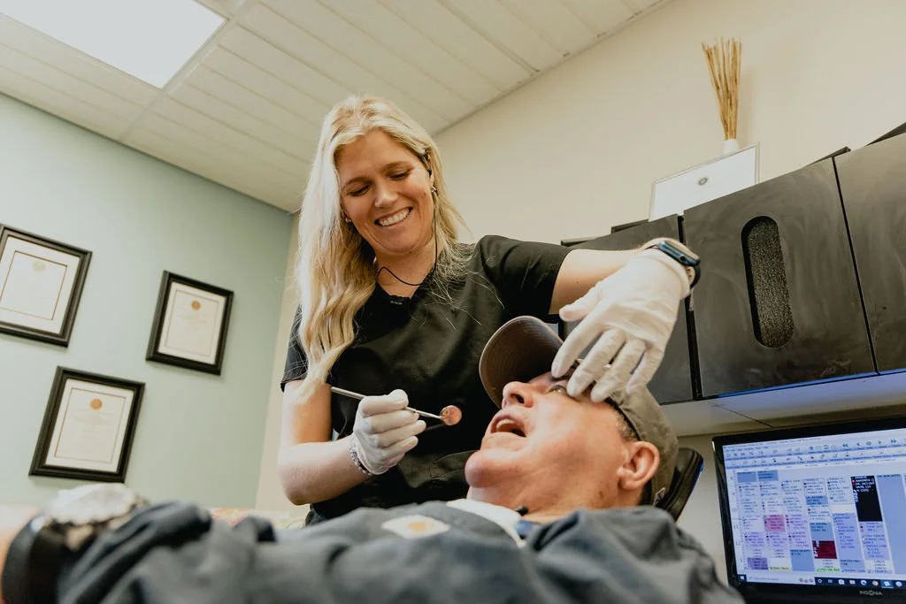 A dentist is performing a dental examination on a patient in a clinic with framed certificates on the wall.