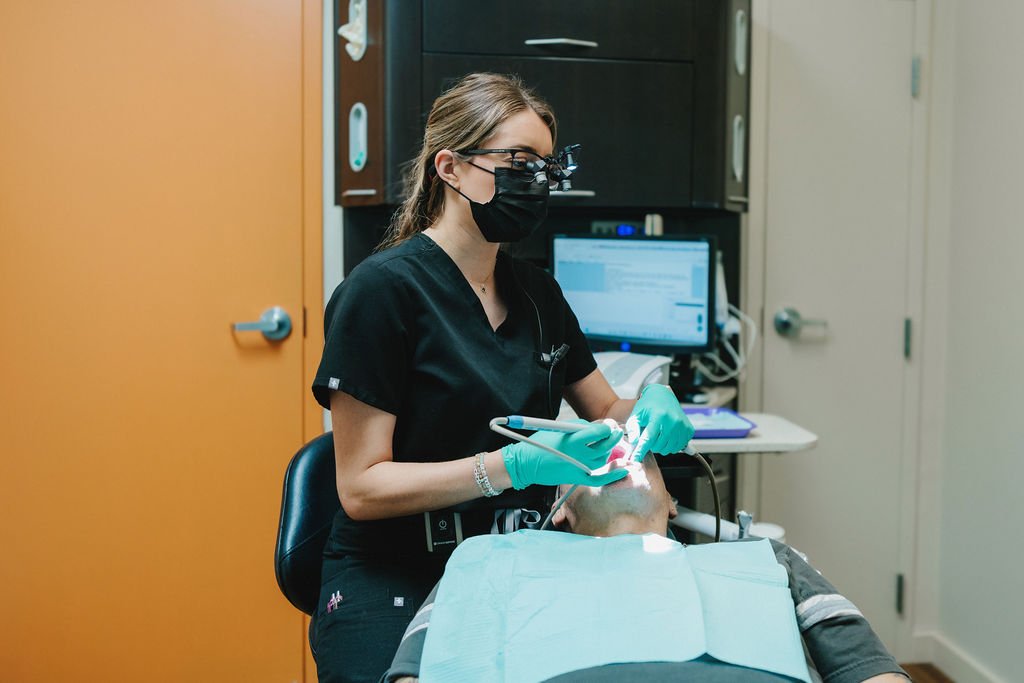 Dentist wearing black scrubs, a mask, and magnifying loupes performing dental work on a patient in a dental clinic.