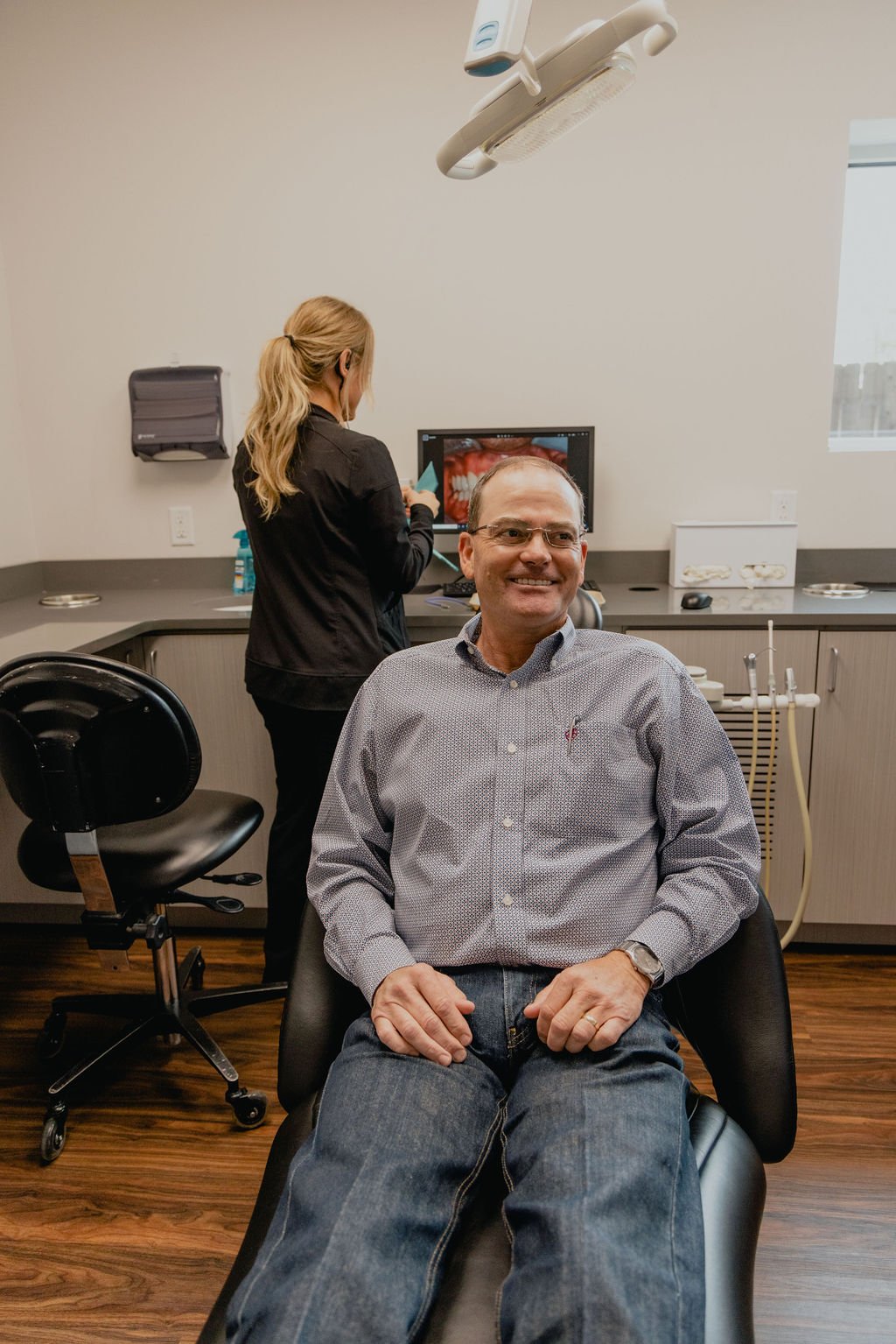 A man sitting in a dentist's chair smiling, with a dental professional in the background looking at a computer screen displaying a close-up of teeth inside the dental office.