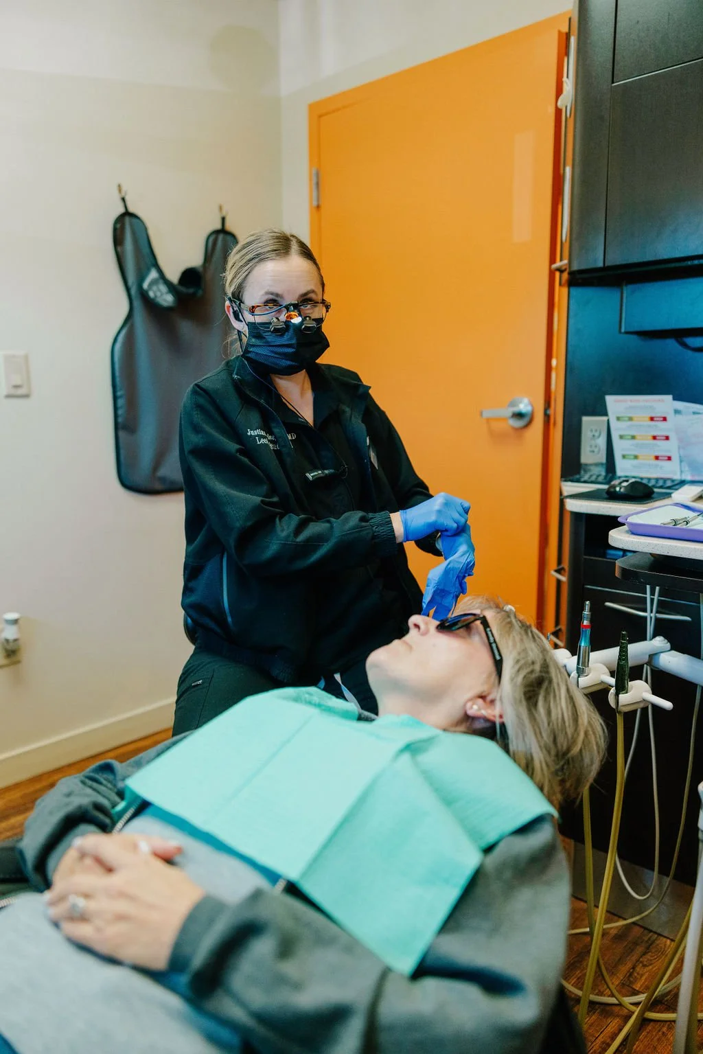 A woman receiving dental treatment from a dentist in a clinic, the dentist wearing a mask, glasses, and gloves, while the patient lies on a dental chair.