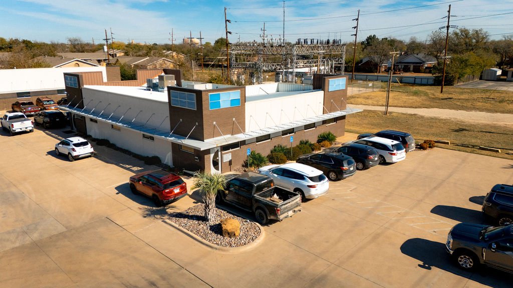 An aerial view of a small commercial building with a parking lot filled with cars, situated in a suburban area with power lines and residential houses in the background.
