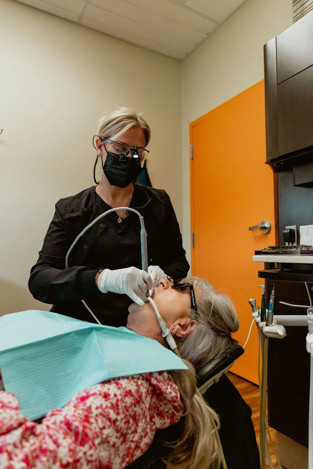 A dentist wearing a face mask, glasses, and gloves performs a dental procedure on a patient reclining in a dental chair. The patient has long gray hair and is wearing sunglasses. The room has an orange door and dental tools nearby.