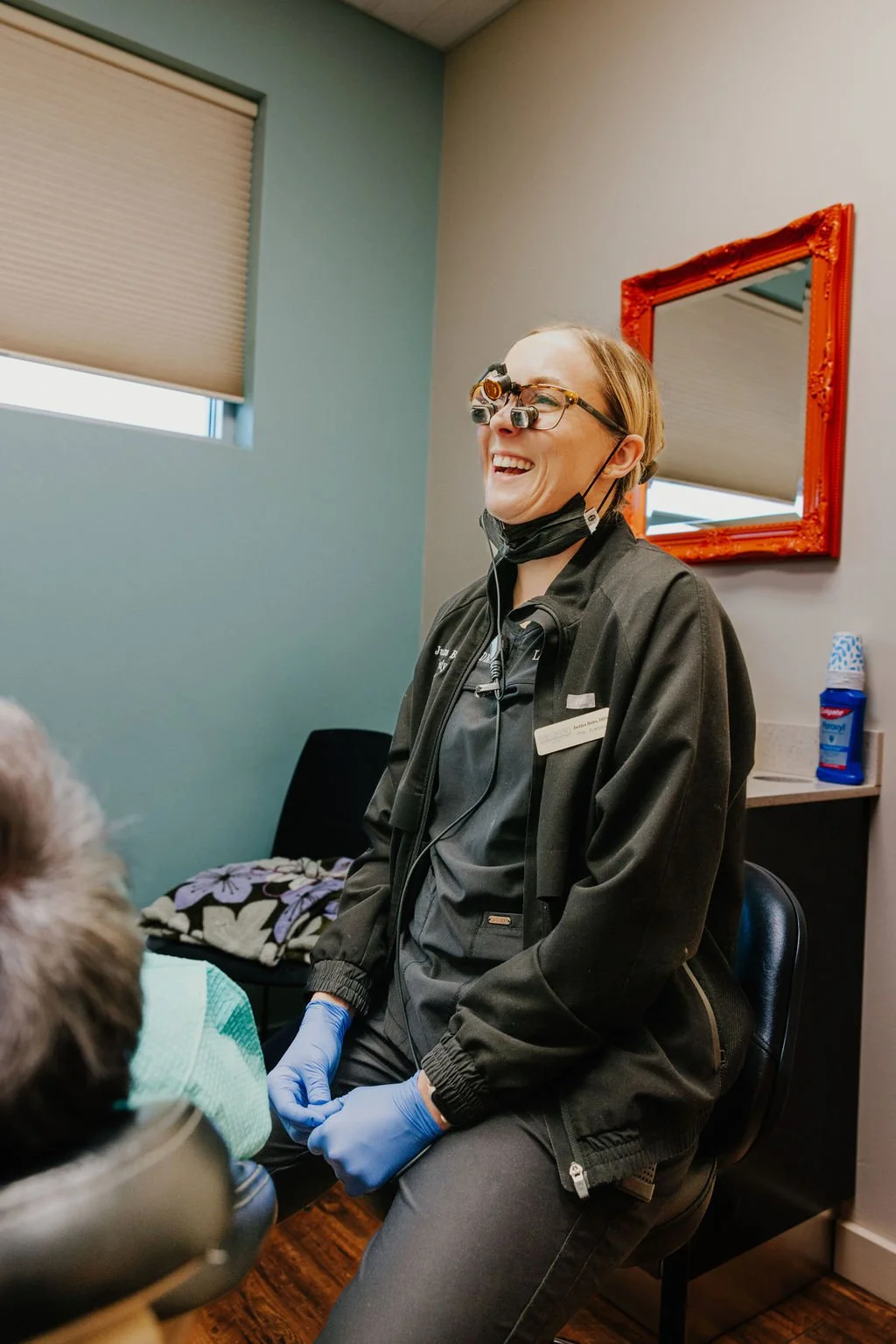 A female dentist or dental professional wearing magnifying loupes and gloves, smiling and talking to a patient in a dental office.