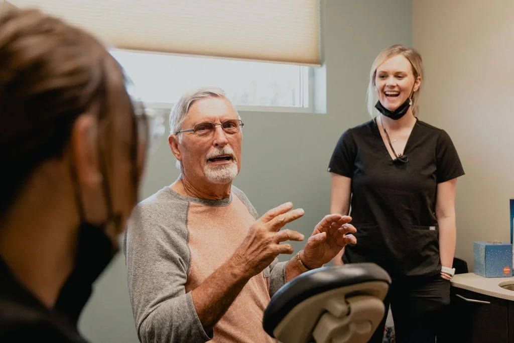An elderly man with gray hair and glasses speaking in a conversation, with a young woman with blonde hair standing nearby and smiling, while a third person with glasses and a black mask is partially visible in the foreground.