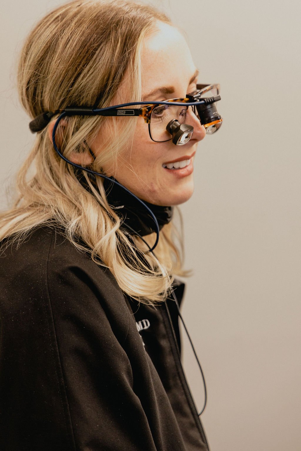 A woman with long blonde hair wearing a black jacket and eye examination glasses with magnifying lenses, smiling and looking to the side.