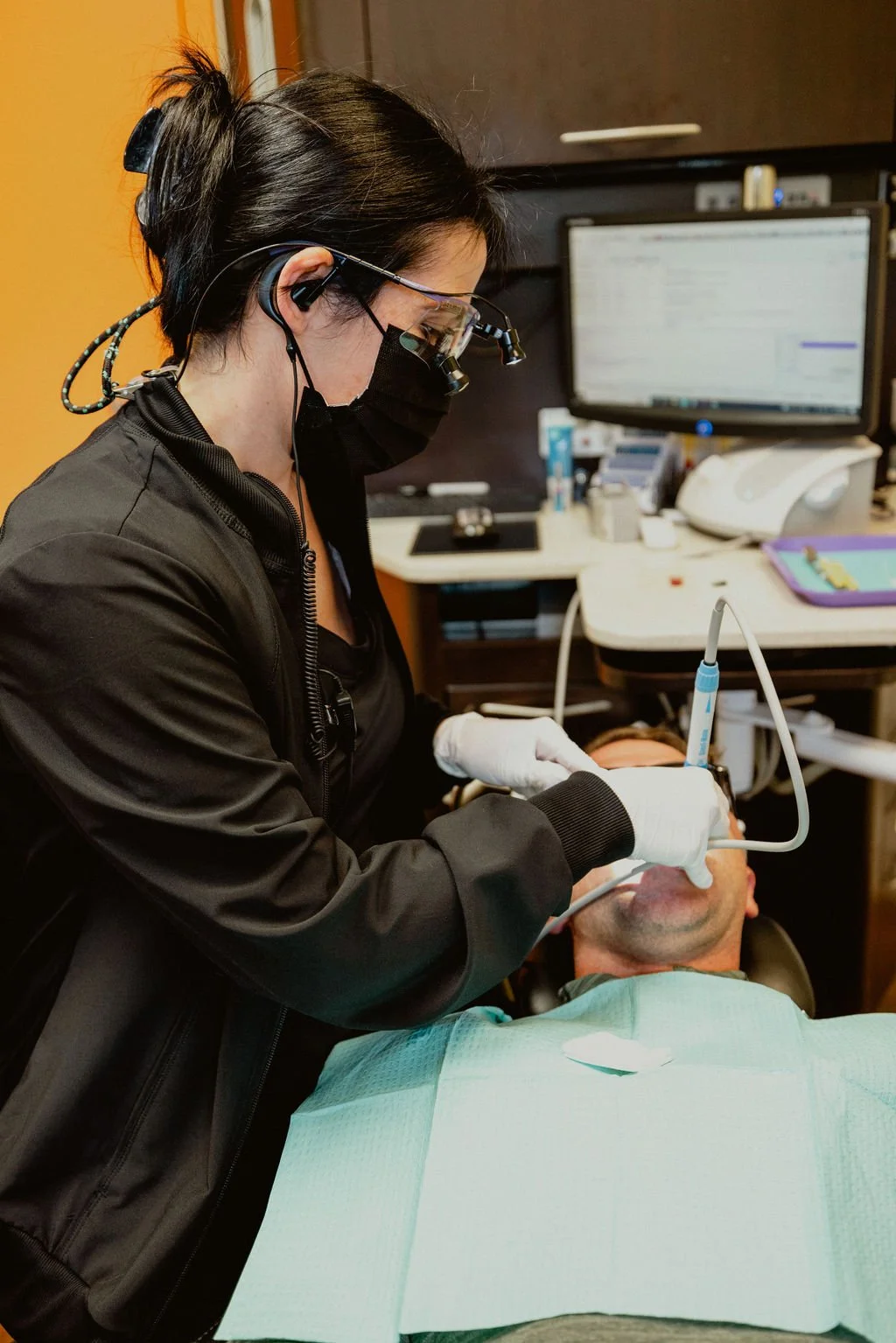 Dental hygienist wearing protective glasses, mask, and gloves performing a dental cleaning on a patient in a dental clinic.