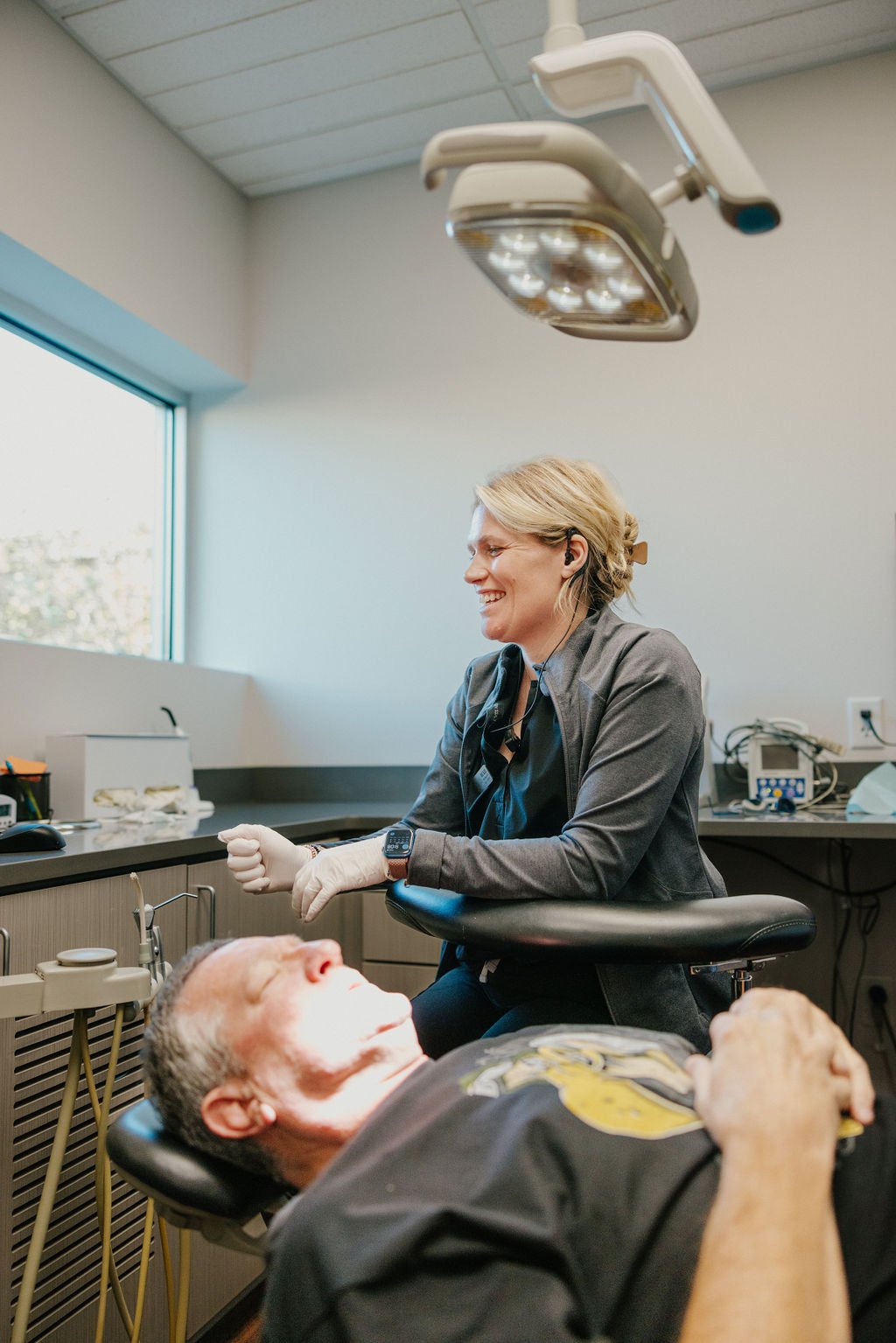 A female healthcare worker with short blonde hair, wearing scrubs and gloves, smiling while talking to a man lying on a medical chair in a clinic or hospital room.