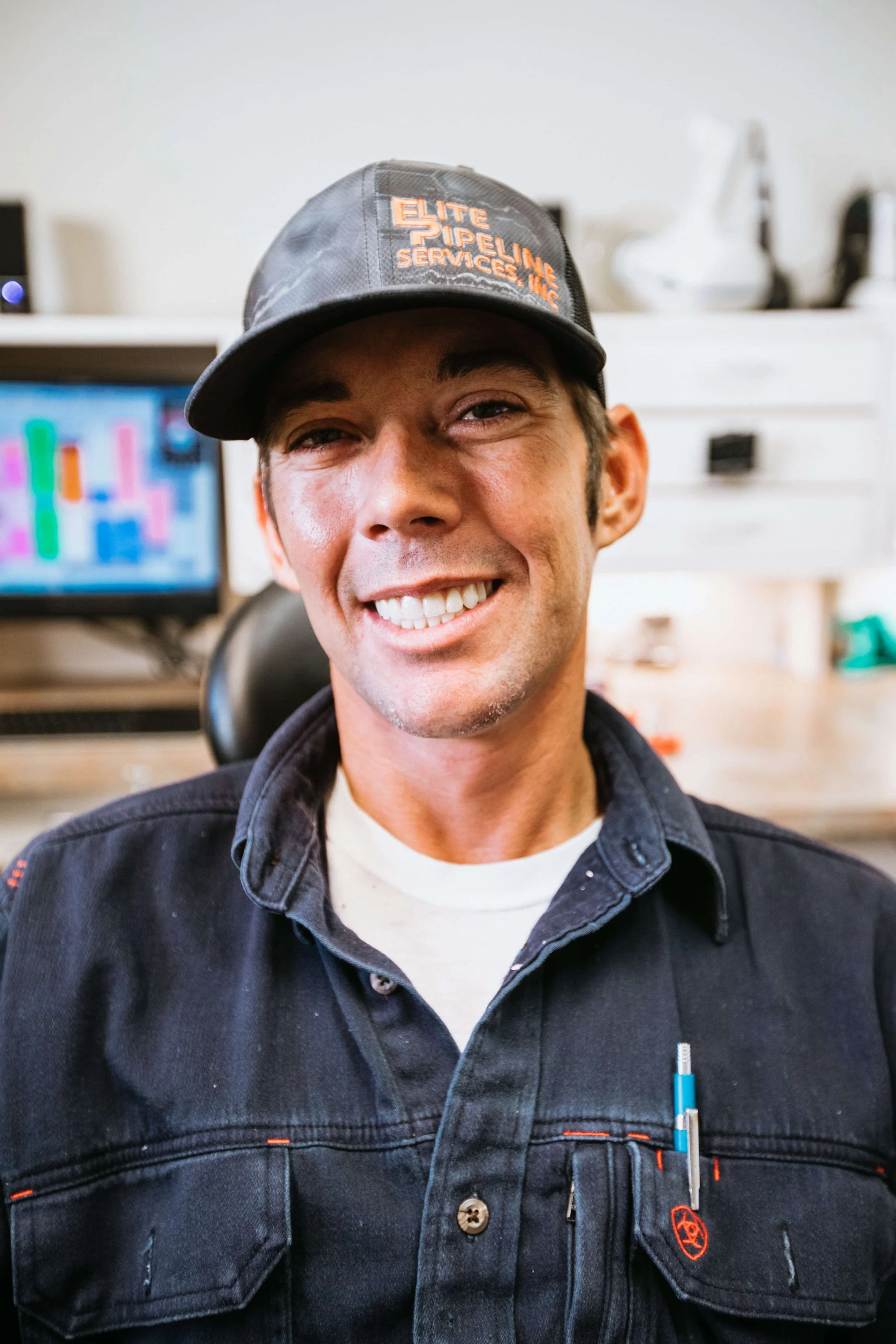 A smiling man wearing a black cap with orange embroidery that reads 'ELITE PIPELINE SERVICES, INC.' and a black work jacket with orange accents, sitting in an office with a computer monitor and white shelf in the background.