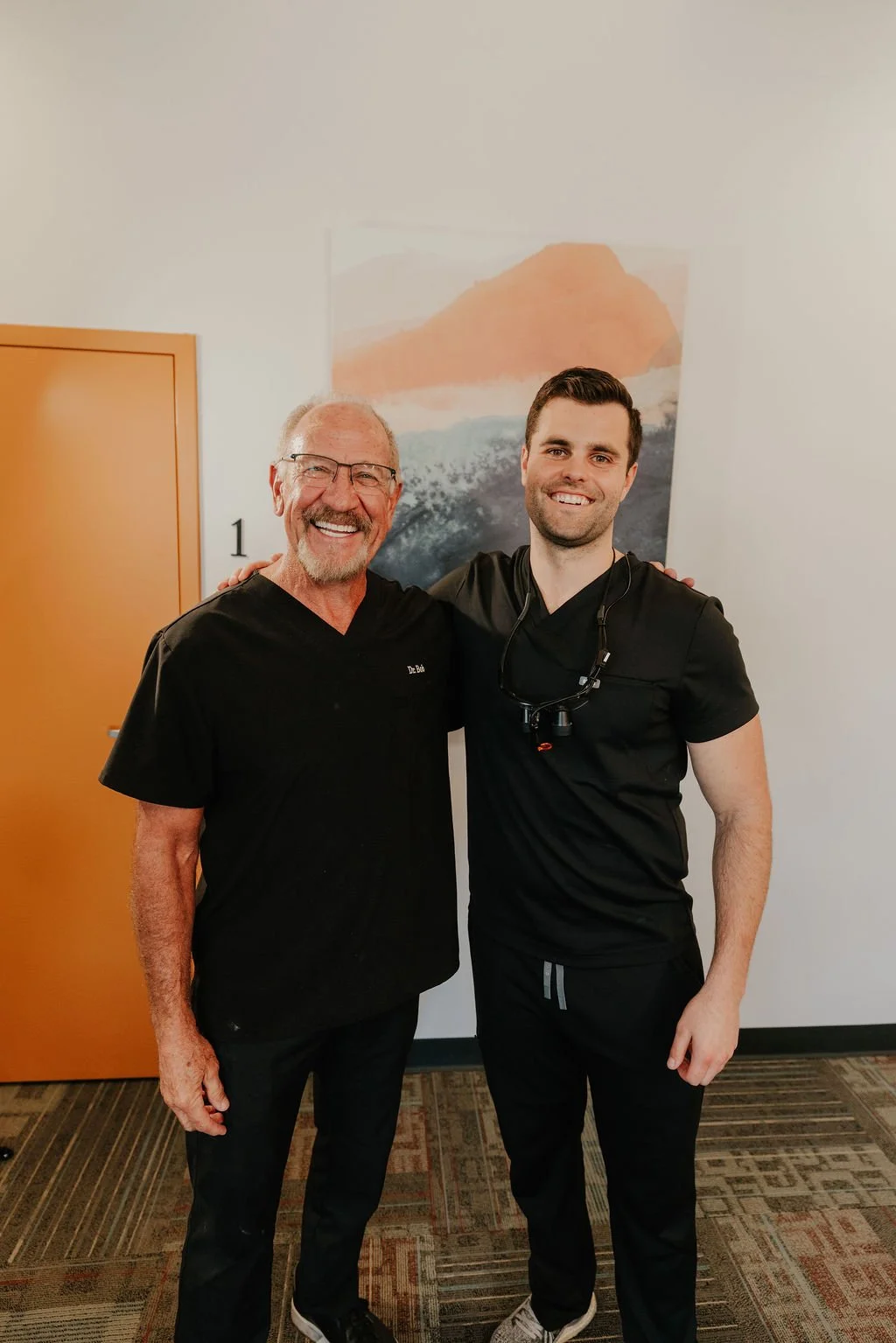 Two men, one older with glasses and a beard, the other younger, standing together indoors. They are smiling and wearing black medical scrubs. The older man has his hand on the younger man's shoulder. A painting of a mountain landscape hangs on the white wall behind them.