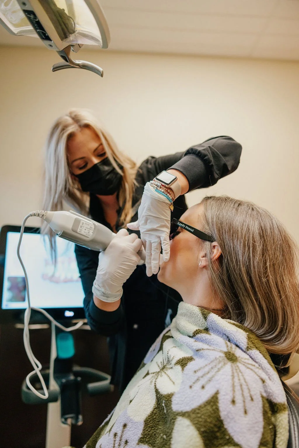 Dental professional examining or treating a patient in a dental office while the patient wears glasses and a towel around her shoulders.