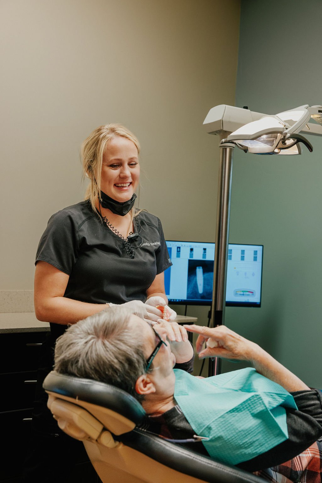 A dental professional smiling and talking with a patient lying in a dental chair. The patient is elderly, wearing glasses, and has a dental bib. There is dental equipment and a monitor displaying dental X-ray images in the background.