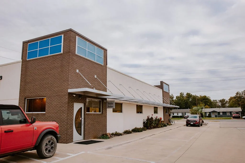 A two-story brick building with a sign that reads 'Smile Suites,' with a parking lot and cars in front, and a partly cloudy sky overhead.