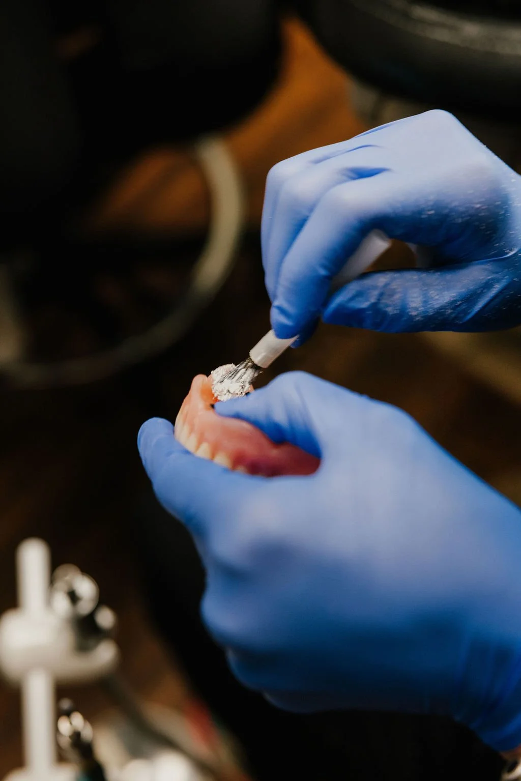 A person wearing blue gloves applying a powdery substance to a set of dentures with a small brush.