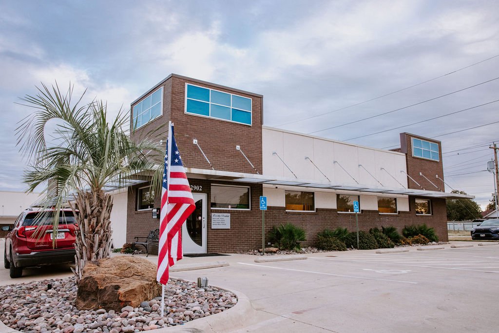 A commercial building with a brick facade, large windows, and a white awning, with parking spaces in front, including designated handicapped spots, and an American flag near a bush and a large rock.