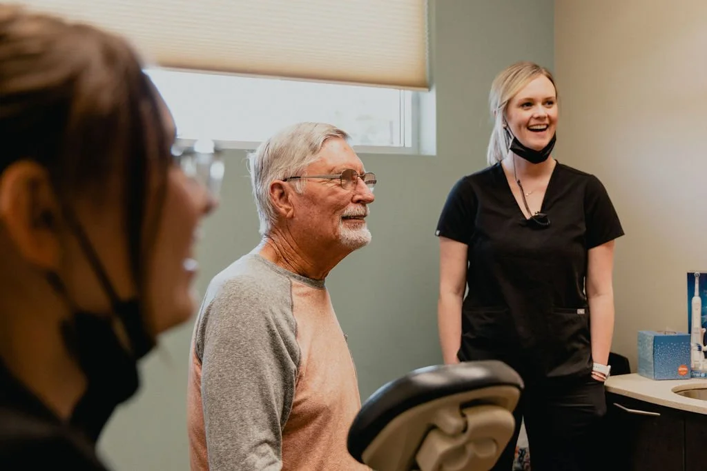 A man at a dental appointment or consultation, smiling and sitting in a dental chair, with a dental professional and another person nearby in a dental office.