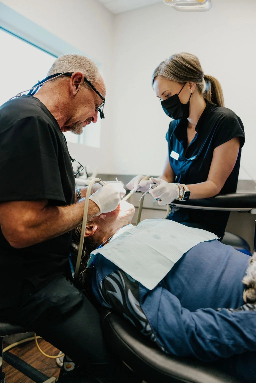 Dentist and dental assistant performing a procedure on a patient in a dental clinic, both wearing black masks and gloves.