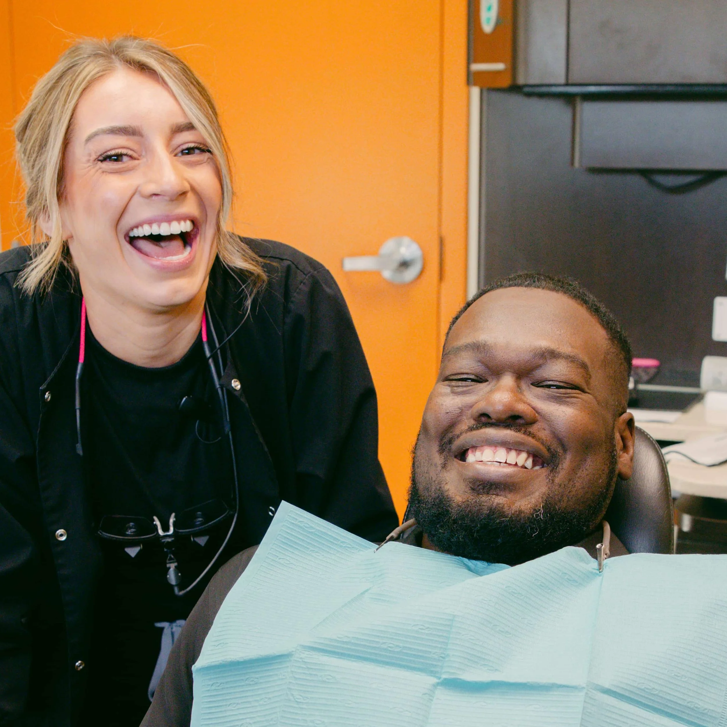 A woman with blonde hair and a wide smile, showing her teeth, is leaning in close to a person with dark skin and a big smile. They are in an indoor setting with orange and gray walls, and there are medical or dental supplies on the desk in front of them.