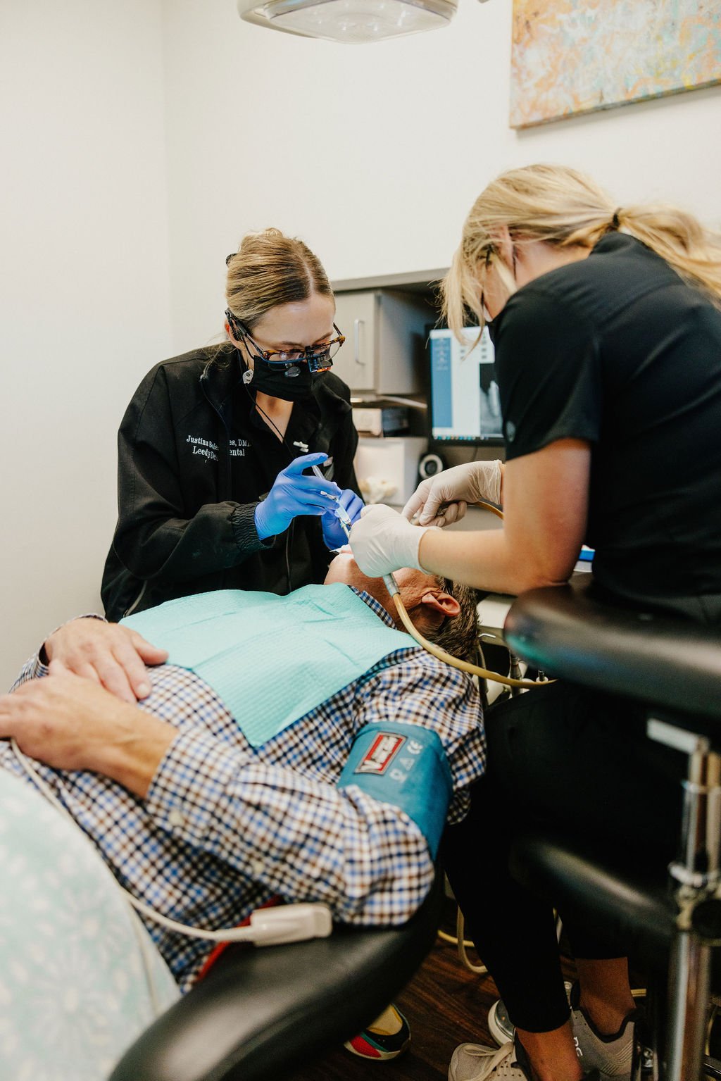 Two dental professionals wearing masks and gloves attending to an elderly patient lying in a dental chair, preparing for a dental procedure in a clinical setting.
