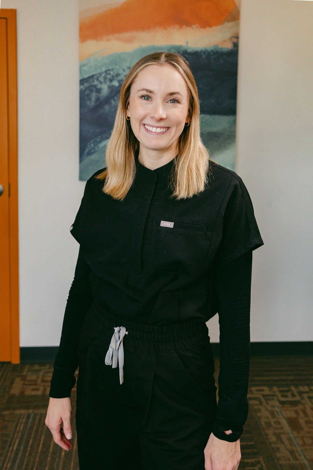 A woman with shoulder-length blonde hair smiling, wearing a black uniform with a badge, standing indoors with a painting of a landscape in the background.