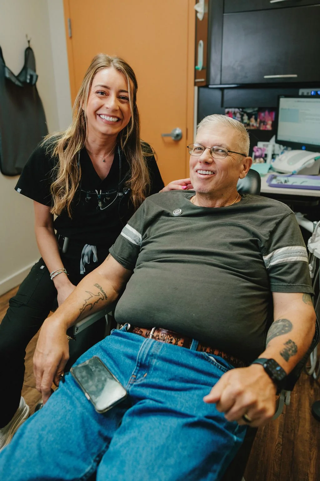 A smiling nurse stands beside a seated man in a medical office. The man is in a hospital chair, wearing a green t-shirt, blue jeans, and glasses. He has tattoos on his arms and a phone on his lap. The background shows medical equipment and office supplies.