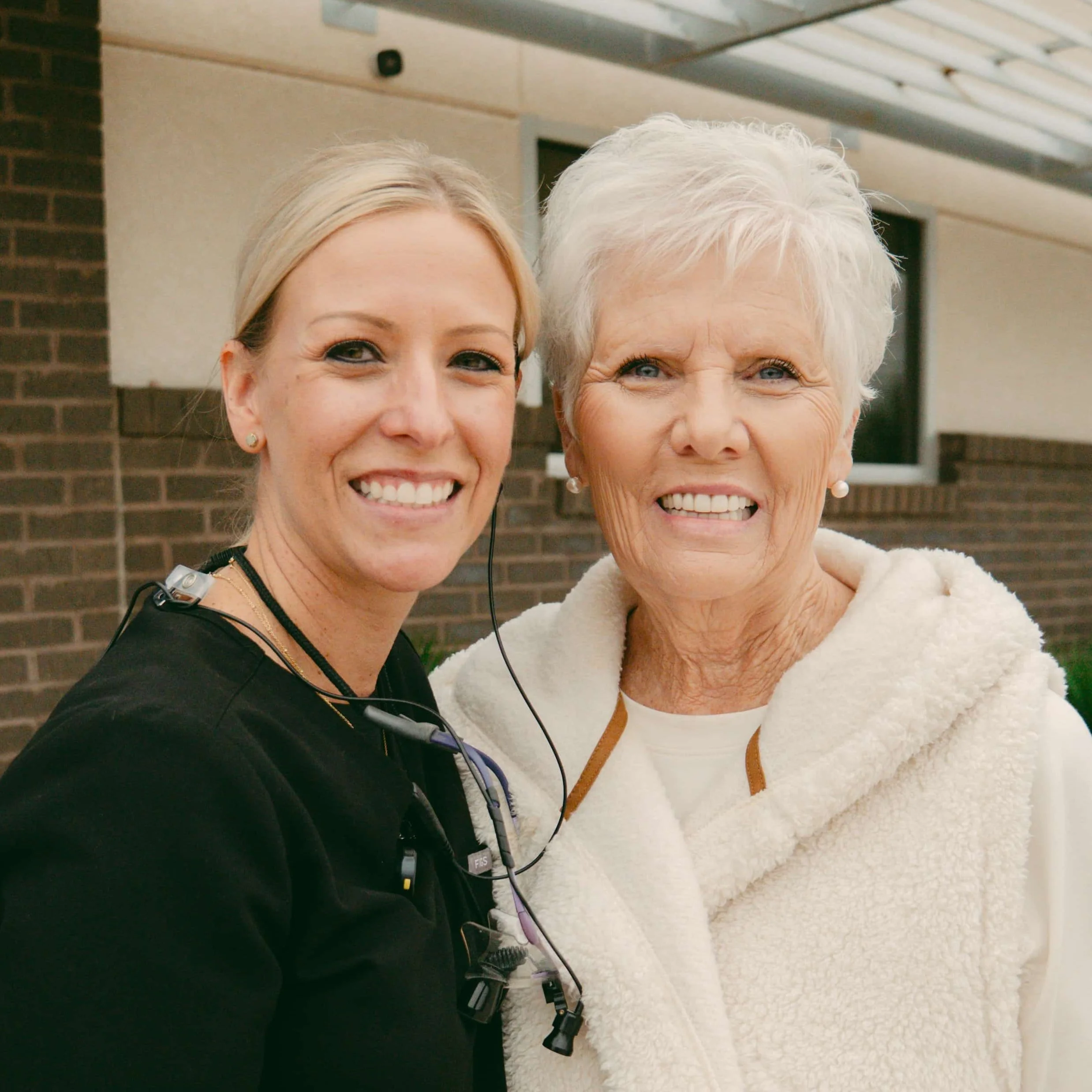 Two women smiling for a selfie, one with blonde hair and the other with white hair, standing outside in front of a brick building. The woman on the right wears a white fuzzy jacket, and the woman on the left wears a black top. Both are wearing earrings and the woman on the left has a microphone and recording device attached to her clothing.