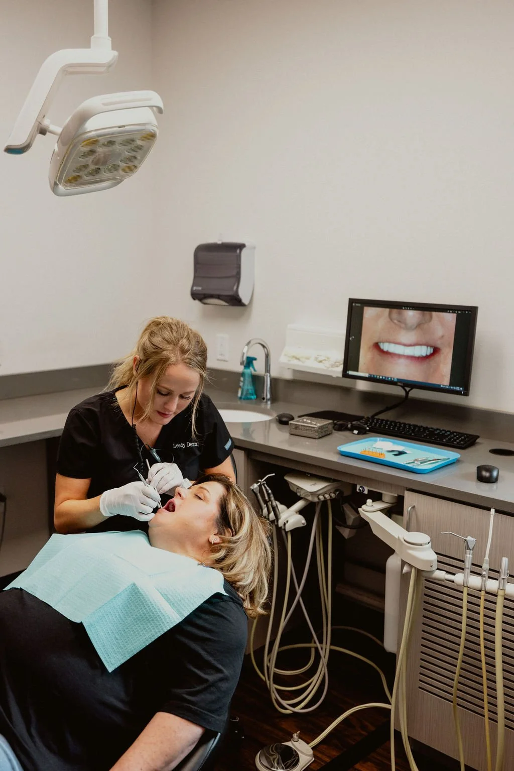 A dental professional examining a patient’s mouth in a dental office, with dental tools and a computer monitor displaying a close-up of a smile.