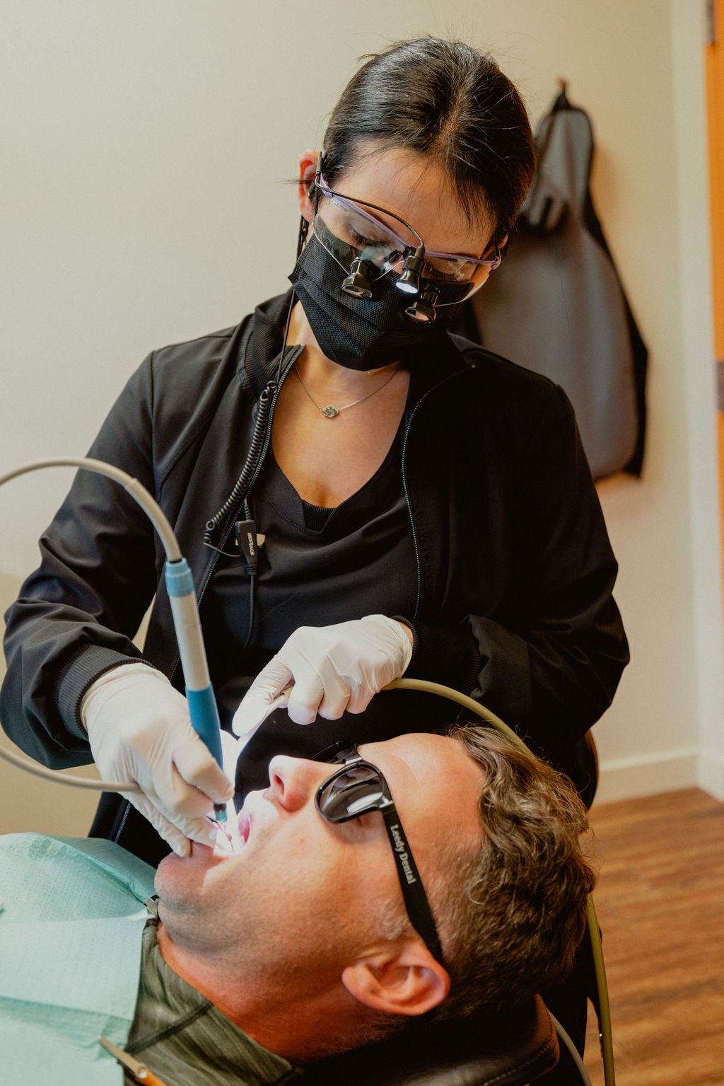 A dentist wearing protective goggles, a mask, and gloves performs a dental procedure on a male patient lying in a dental chair with sunglasses on.