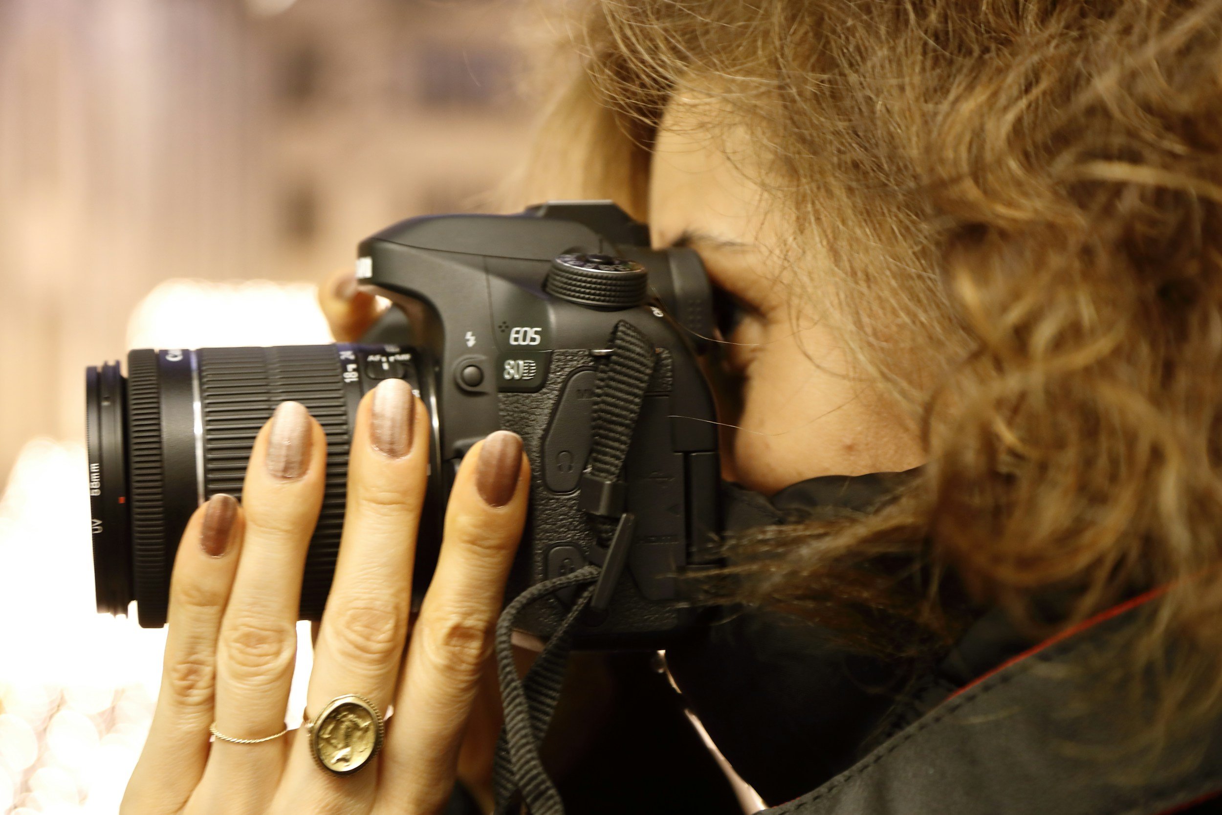 A woman with curly hair holding a DSLR camera up to her eye, taking a photograph.