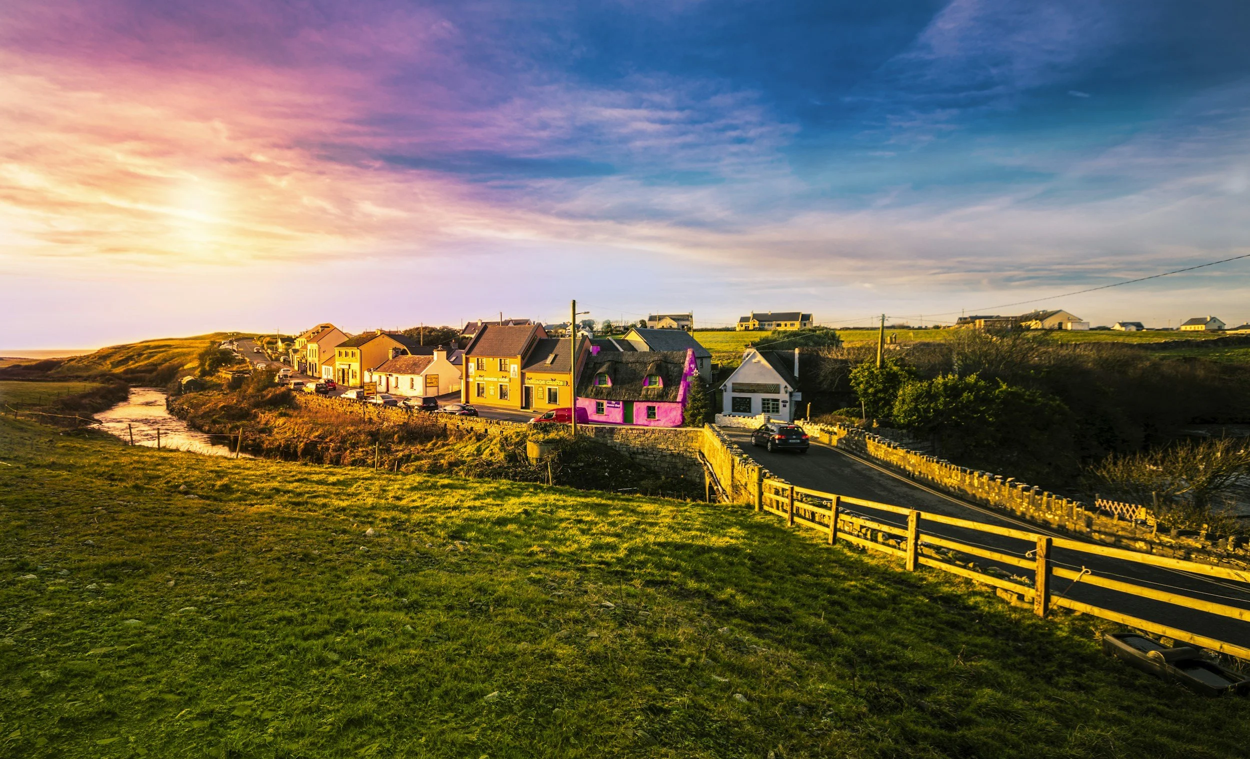 Colorful houses along a winding road with a grassy foreground and a sunset sky.