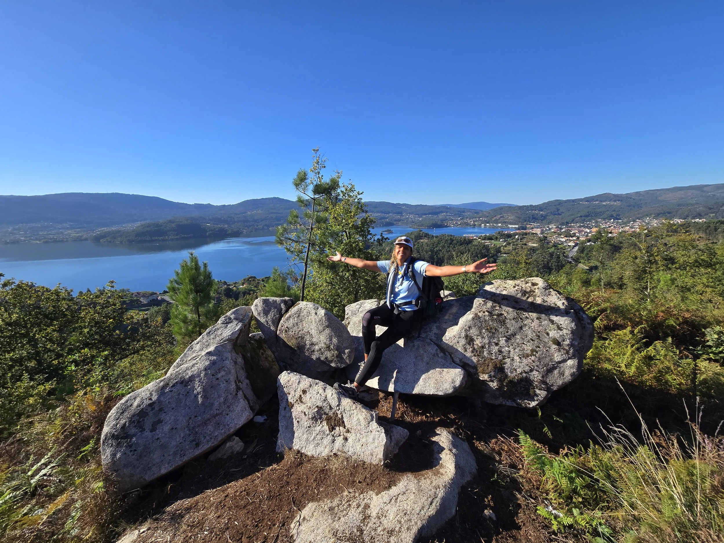 Hiker with outstretched arms sitting on large rocks on a hilltop overlooking a lake, green forests, and mountains under a clear blue sky.