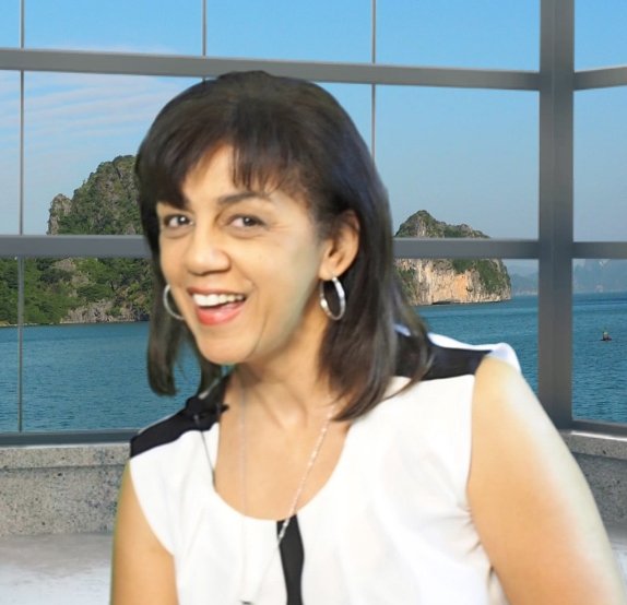 A woman with shoulder-length dark hair wearing hoop earrings and a white sleeveless top, smiling at the camera, with a large window showing an ocean view and islands in the background.