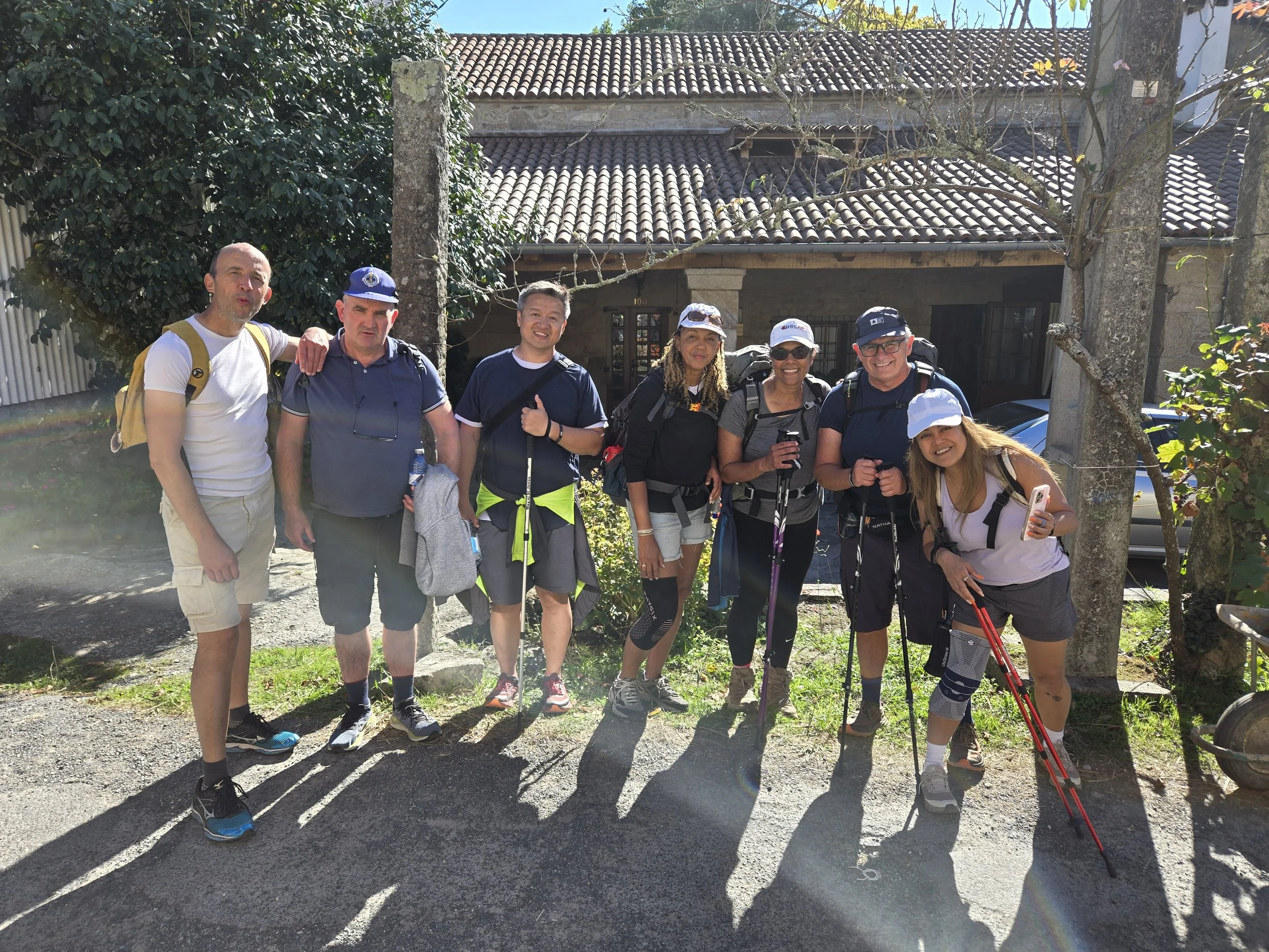 Group of eight hikers standing outdoors on a sunny day, some holding trekking poles, in front of a house with a tiled roof and trees.