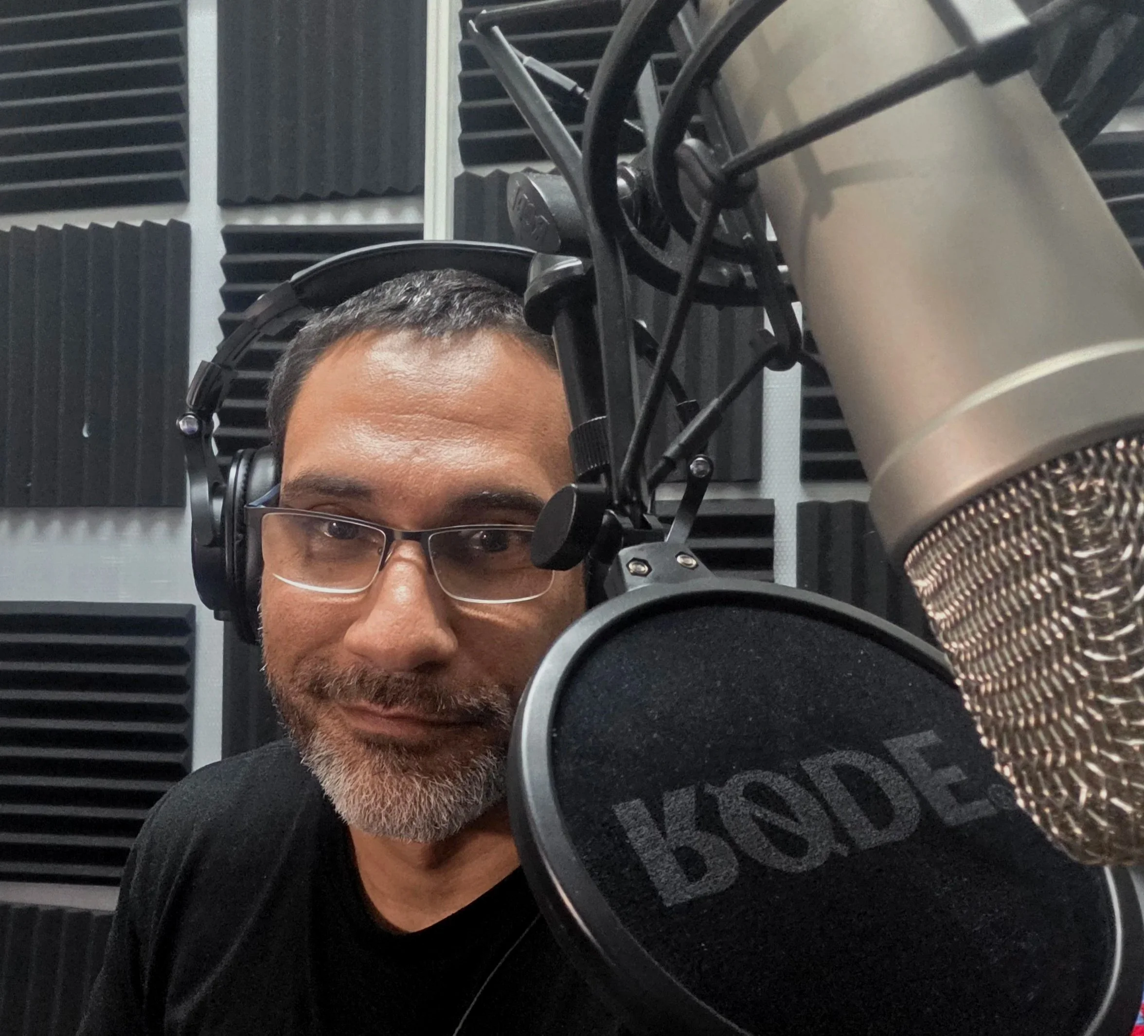 A man wearing glasses and headphones in a recording studio, positioned behind a microphone labeled 'BOSE', with black soundproofing foam on the walls.